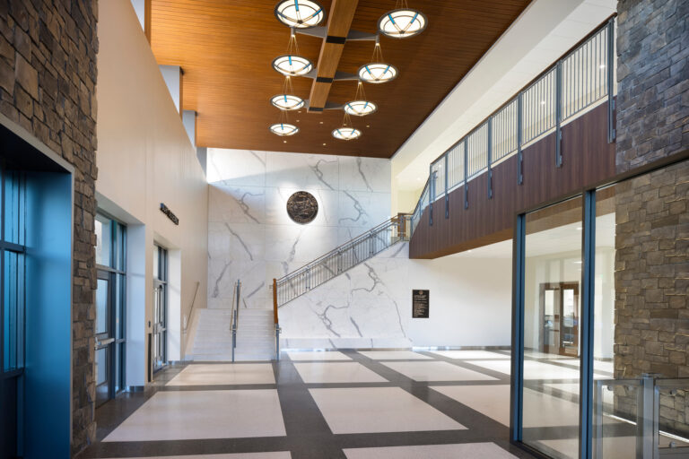 Modern civic building lobby with stone walls, marble staircase, wood ceiling, and circular pendant lights.