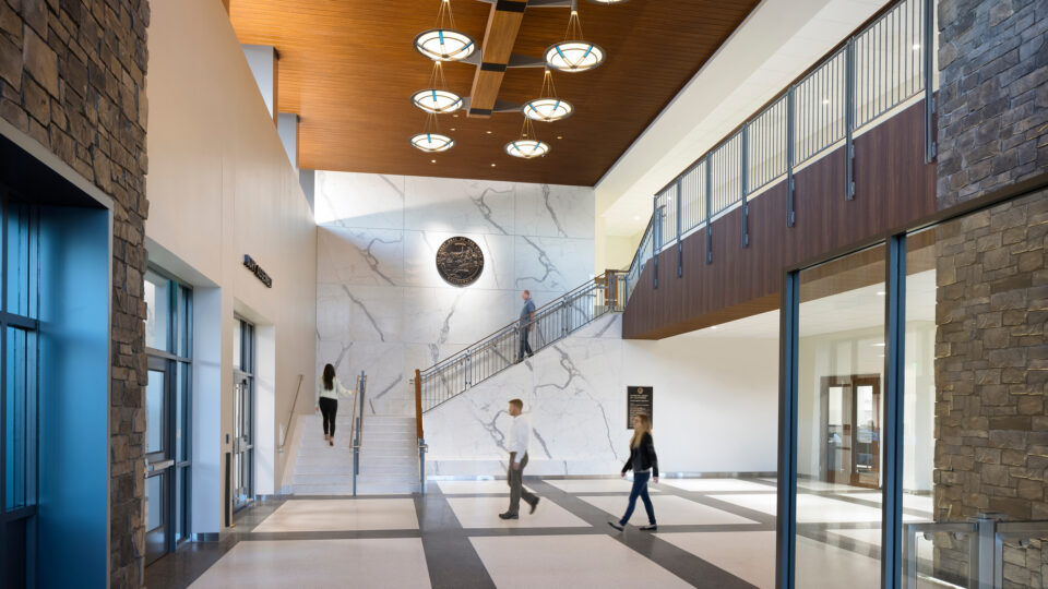 Spacious modern courthouse lobby with stone walls, a marble stairway, wood ceiling, and people walking through.