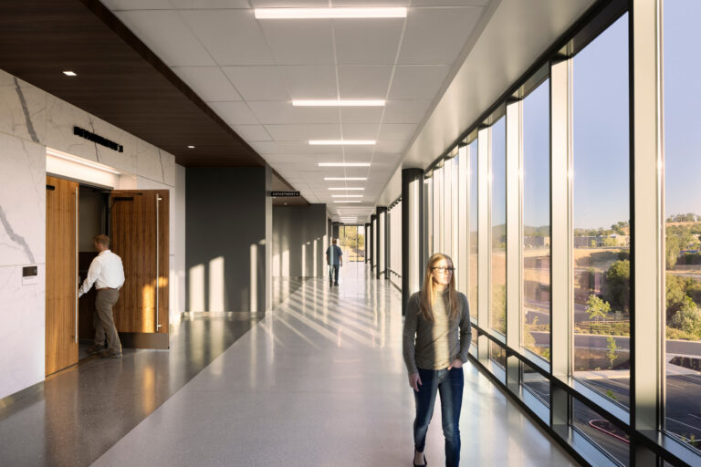 Sunlit modern hallway with floor-to-ceiling windows, a woman walking, and a man entering a room on the left