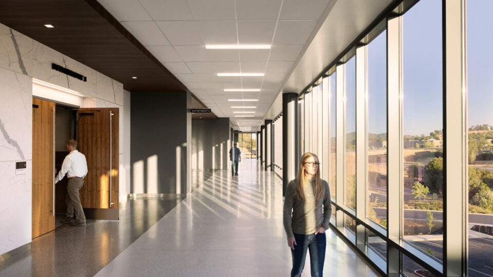 Sunlit modern hallway with floor-to-ceiling windows, a woman walking, and a man entering a room on the left