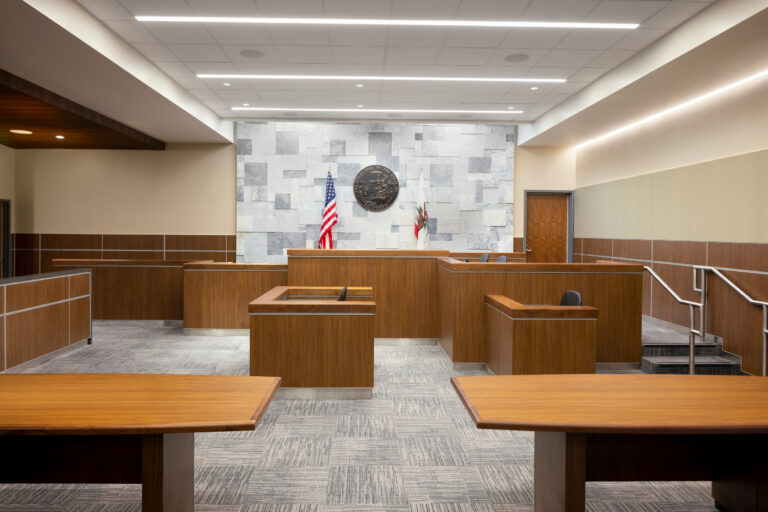 Modern courtroom interior with wood benches, witness stand, judge’s bench, and U.S. and California flags.