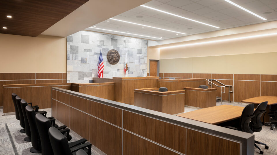 Modern courtroom interior with wood paneling, judge’s bench, jury box, and U.S. and California flags beneath the state seal