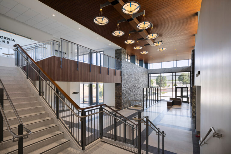 Spacious modern building lobby with staircases, metal railings, stone walls, large windows, and circular ceiling lights