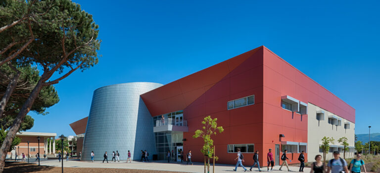Students walking past a modern red and white campus building with a silver cylindrical entrance under a clear blue sky
