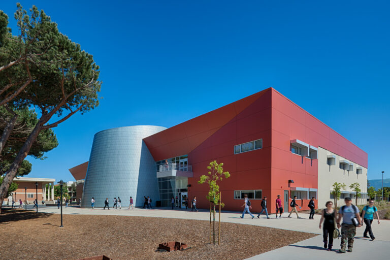 Students walking past a modern red and white campus building with a silver cylindrical entrance under a clear blue sky