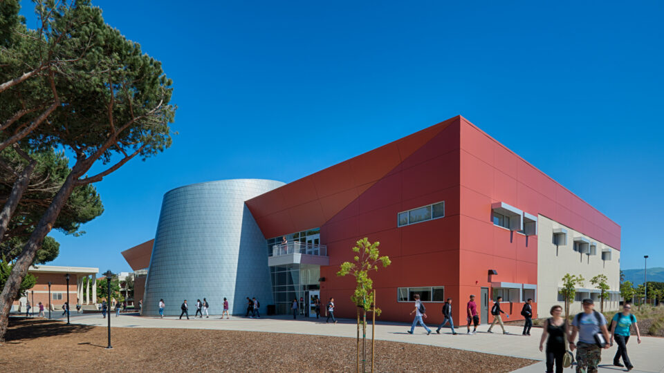Students walking past a modern red and white campus building with a silver cylindrical entrance under a clear blue sky