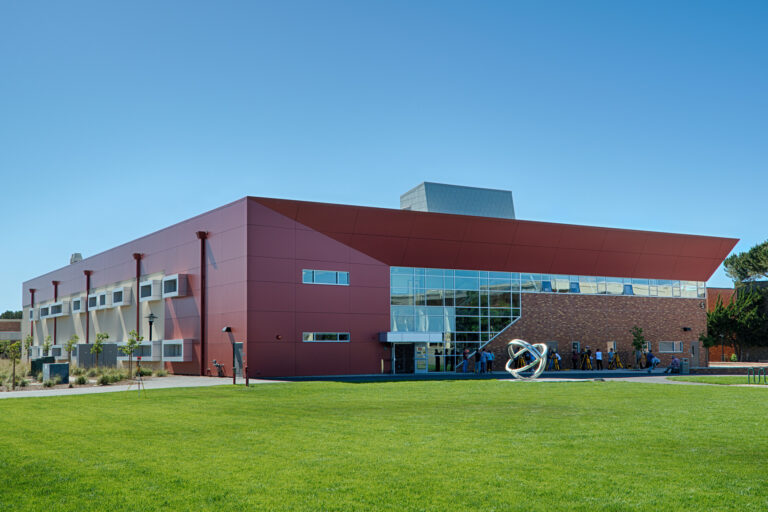 Modern red and brick campus building with large glass windows, a sculpture outside, and a wide green lawn under clear sky