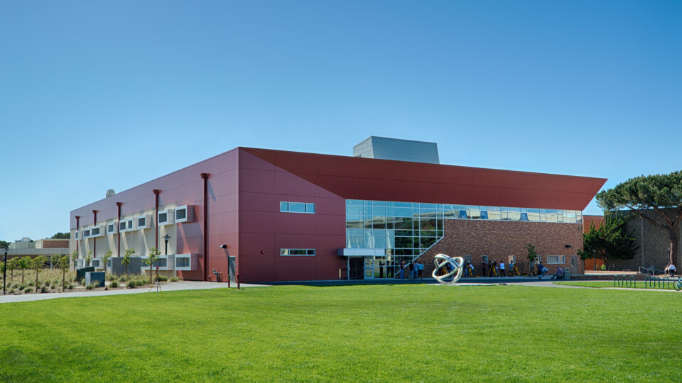 Modern red and brick campus building with large glass windows, a sculpture outside, and a wide green lawn under clear sky