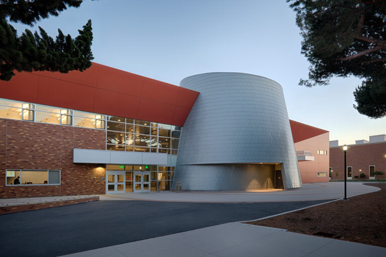Modern brick building with a curved silver facade and red roofline at dusk, framed by tall trees.