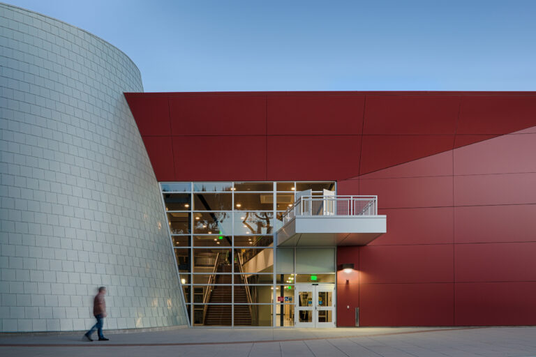 Modern building entrance with a curved white tiled wall, red facade, glass stairwell, and a blurred person walking past