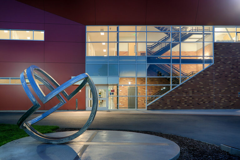 Abstract metal loop sculpture outside a modern building with large lit windows and a visible interior staircase