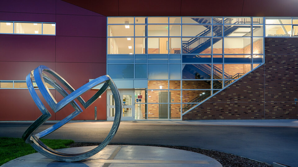 Abstract metal loop sculpture outside a modern building with large lit windows and a visible interior staircase