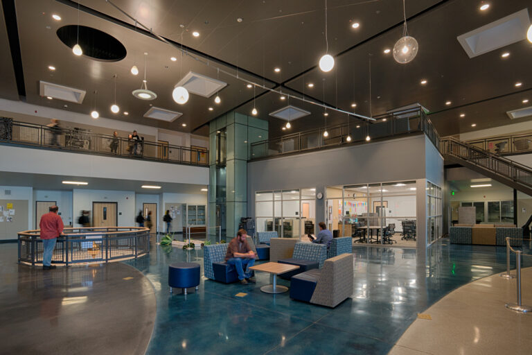 Modern two-story atrium with a glass elevator, staircase, lounge seating, and classrooms or offices around the lobby
