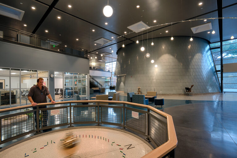 Man standing by a large pendulum exhibit in a spacious modern atrium with hanging lights and glass-walled rooms