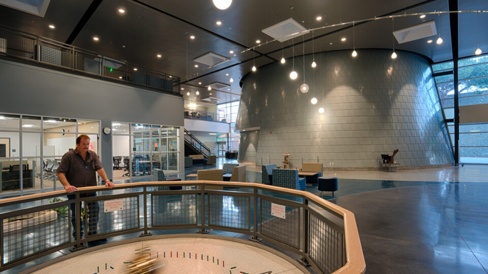 Man standing by a large pendulum exhibit in a spacious modern atrium with hanging lights and glass-walled rooms