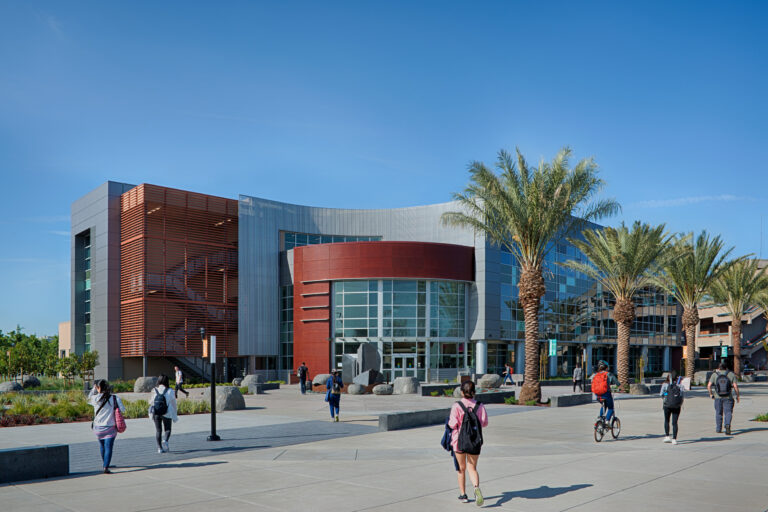 Students walking past a modern campus building with glass walls and palm trees under a clear blue sky