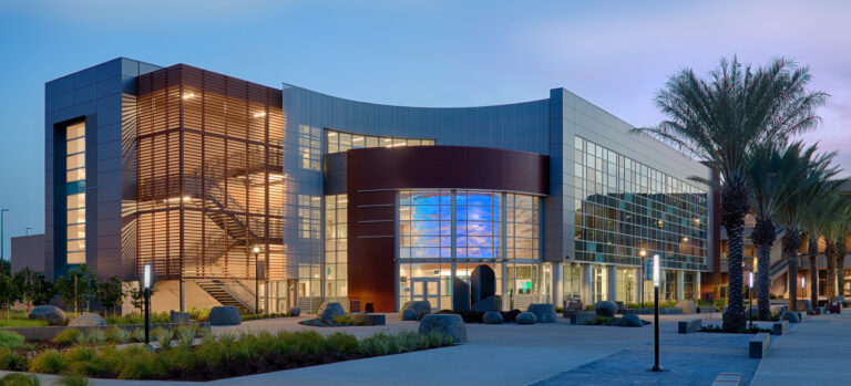 Modern glass-and-metal building at dusk, with a curved facade, lit interior stairwell, and palm-lined courtyard.
