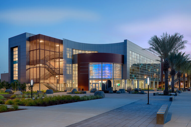 Modern glass-and-metal building at dusk, with a curved facade, lit interior stairwell, and palm-lined courtyard.