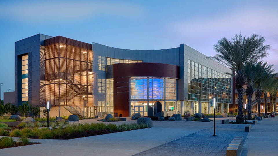 Modern glass-and-metal building at dusk, with a curved facade, lit interior stairwell, and palm-lined courtyard.