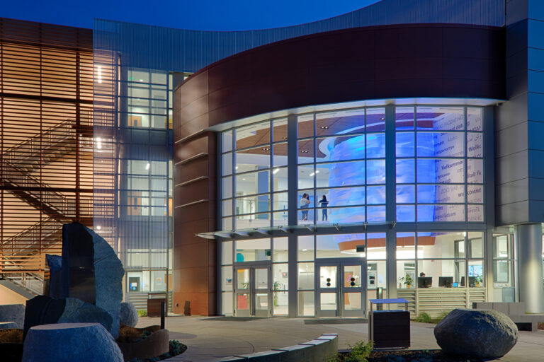 Modern glass-fronted building entrance at dusk with a blue-lit interior feature and welcome text in many languages