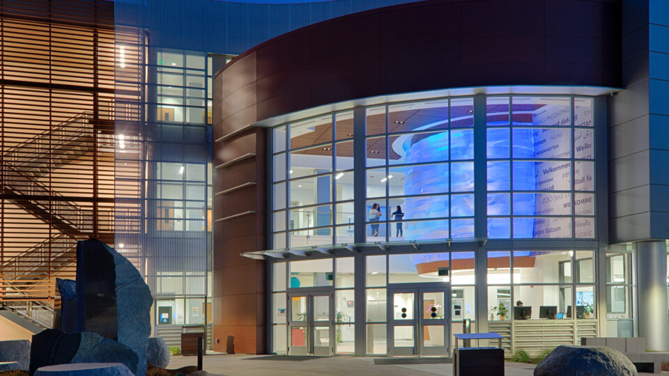 Modern glass-fronted building entrance at dusk with a blue-lit interior feature and welcome text in many languages