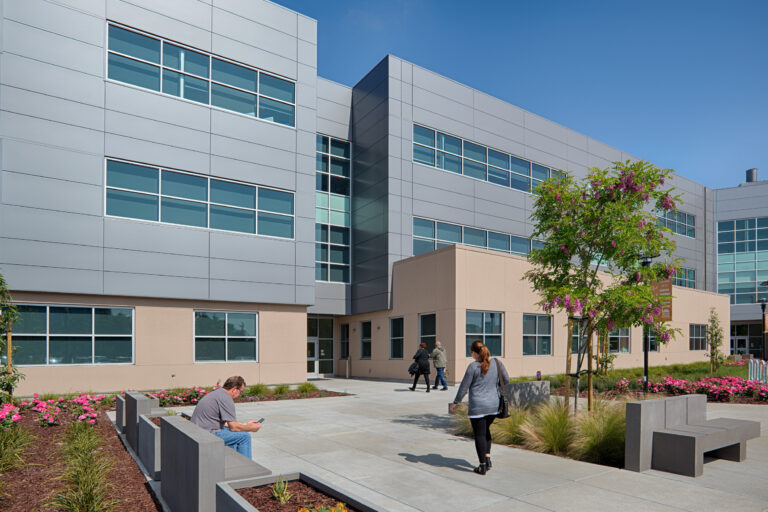 Modern office building with large windows, landscaped courtyard, and people walking and sitting outside.