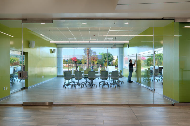 Glass-walled conference room with green walls, rolling chairs, and a person writing on a whiteboard.