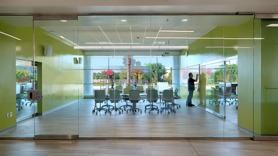 Glass-walled conference room with green walls, rolling chairs, and a person writing on a whiteboard.