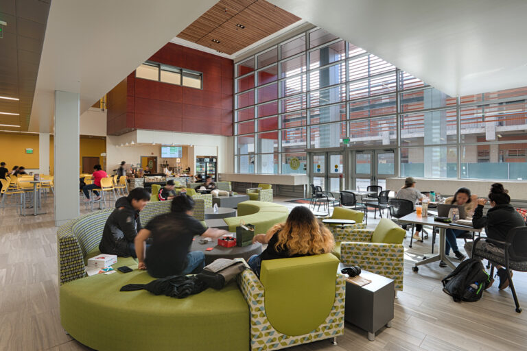 Students gather in a bright modern campus lounge with green seating, tables, and a small cafe by tall windows.