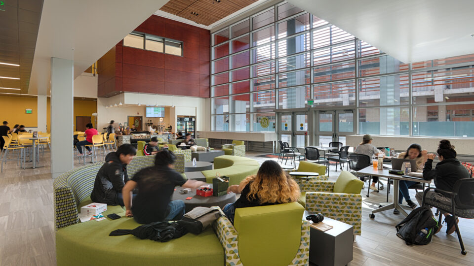 Students gather in a bright modern campus lounge with green seating, tables, and a small cafe by tall windows.