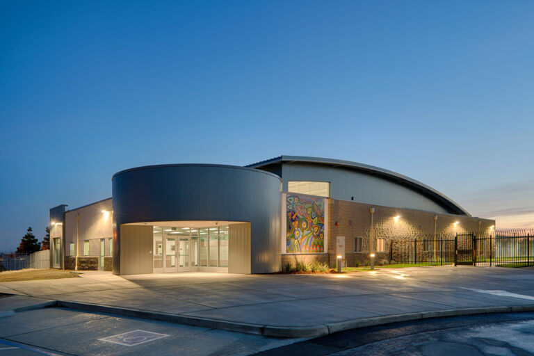 Modern curved-roof building at dusk with a lit glass entrance, mural panel, and fenced perimeter.