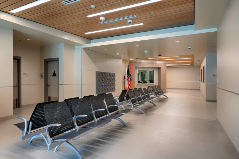 Modern public waiting area with rows of black chairs, restroom doors, lockers, and a service window with flags nearby