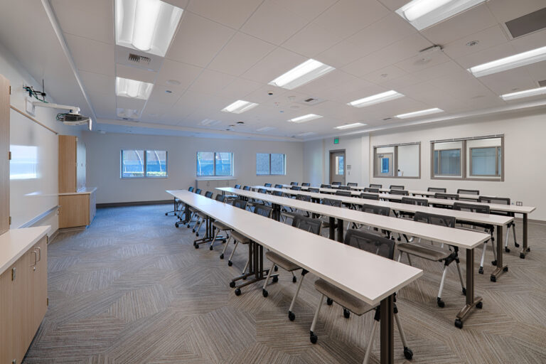 Modern classroom with rows of white tables and gray chairs, a projector, whiteboard, and interior windows.