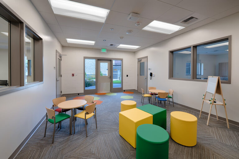 Bright children's activity room with small tables, colorful stools, a whiteboard easel, and glass doors and windows