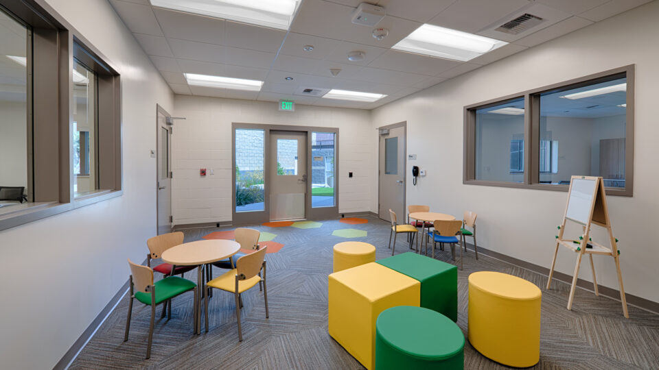 Bright children's activity room with small tables, colorful stools, a whiteboard easel, and glass doors and windows