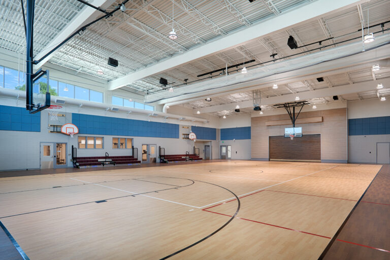 Bright indoor basketball gym with polished wood courts, multiple hoops, high ceilings, and small bleachers.
