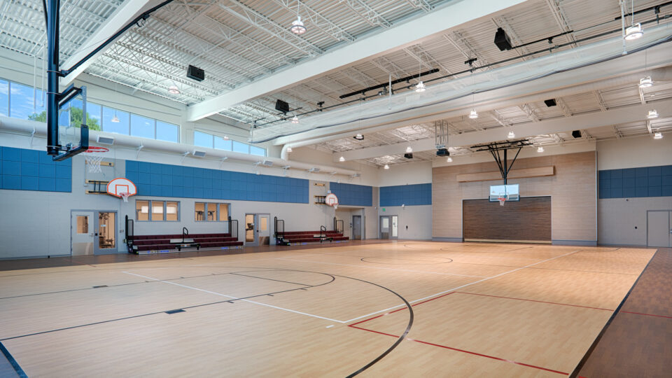 Bright indoor basketball gym with polished wood courts, multiple hoops, high ceilings, and small bleachers.