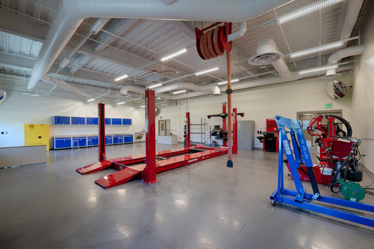 Bright automotive workshop with red vehicle lift, alignment equipment, blue cabinets, and exposed ceiling ducts