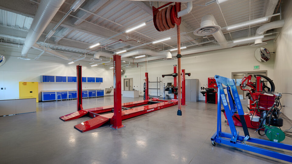 Bright automotive workshop with red vehicle lift, alignment equipment, blue cabinets, and exposed ceiling ducts