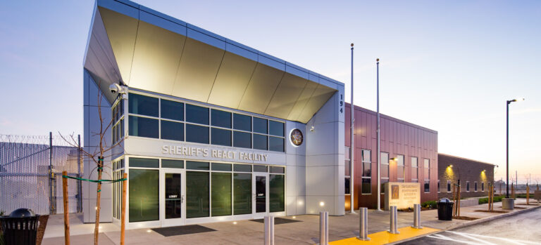 Modern sheriff’s react facility building at dusk with glass entrance, flagpoles, and secure fencing.