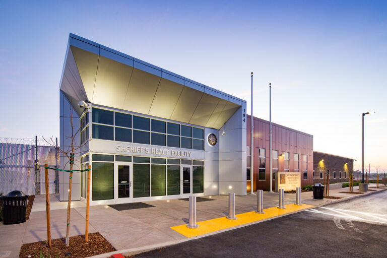 Modern sheriff’s react facility building at dusk with glass entrance, flagpoles, and secure fencing.
