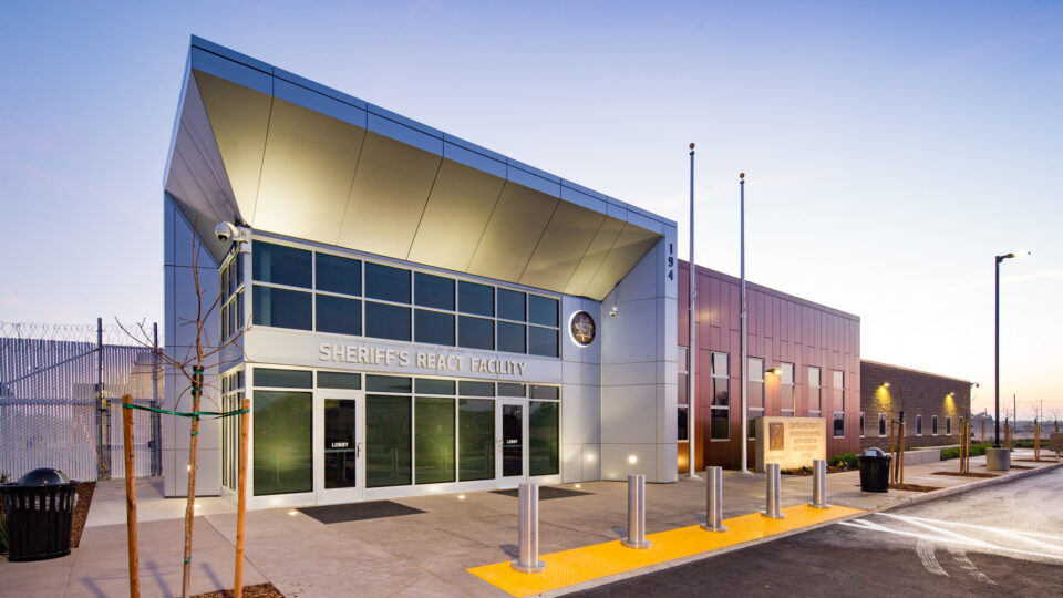 Modern sheriff’s react facility building at dusk with glass entrance, flagpoles, and secure fencing.