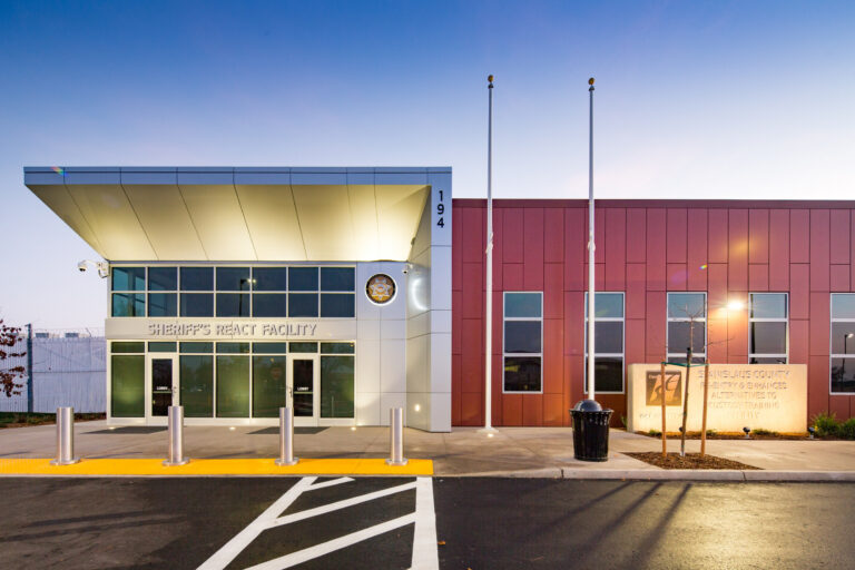 Front entrance of the Sheriff’s REACT Facility at dusk, with modern red and white exterior and flagpoles.