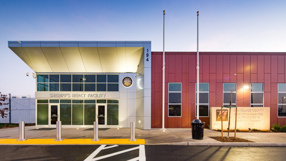 Front entrance of the Sheriff’s REACT Facility at dusk, with modern red and white exterior and flagpoles.