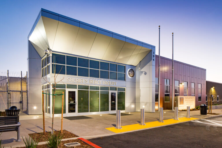 Modern sheriff’s react facility building at dusk with a glass entrance, flagpoles, and security fencing.