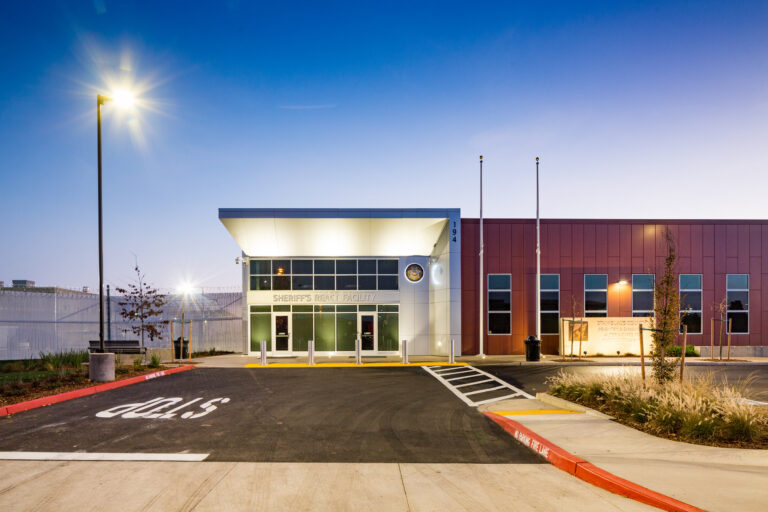 Front entrance of a sheriff’s REACT facility at dusk, with lit windows, flagpoles, and a drive marked CUC.