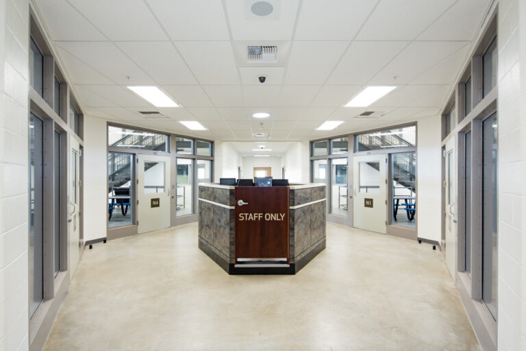 Bright institutional corridor with a central staff-only security desk, glass doors, and numbered room entrances on both sides