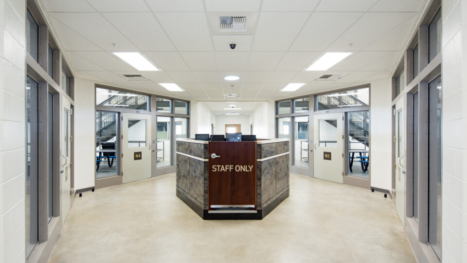 Bright institutional corridor with a central staff-only security desk, glass doors, and numbered room entrances on both sides