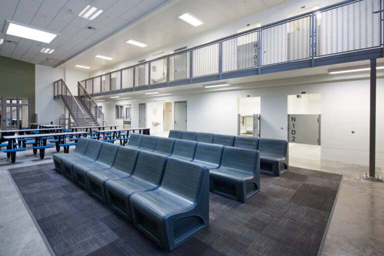 Modern detention facility common area with blue benches, cafeteria tables, upstairs cells, and metal stairway