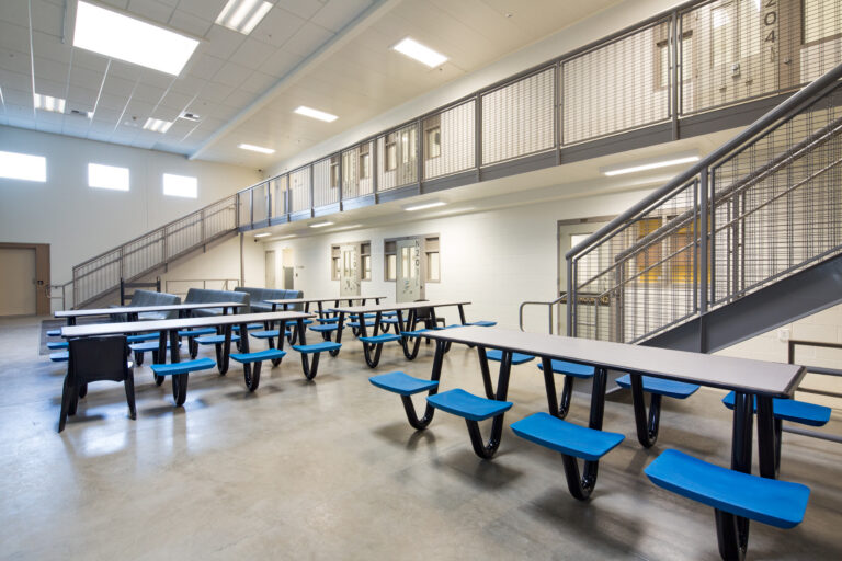Empty institutional common area with cafeteria tables, blue seats, metal staircases, and a second-floor walkway with cell doors